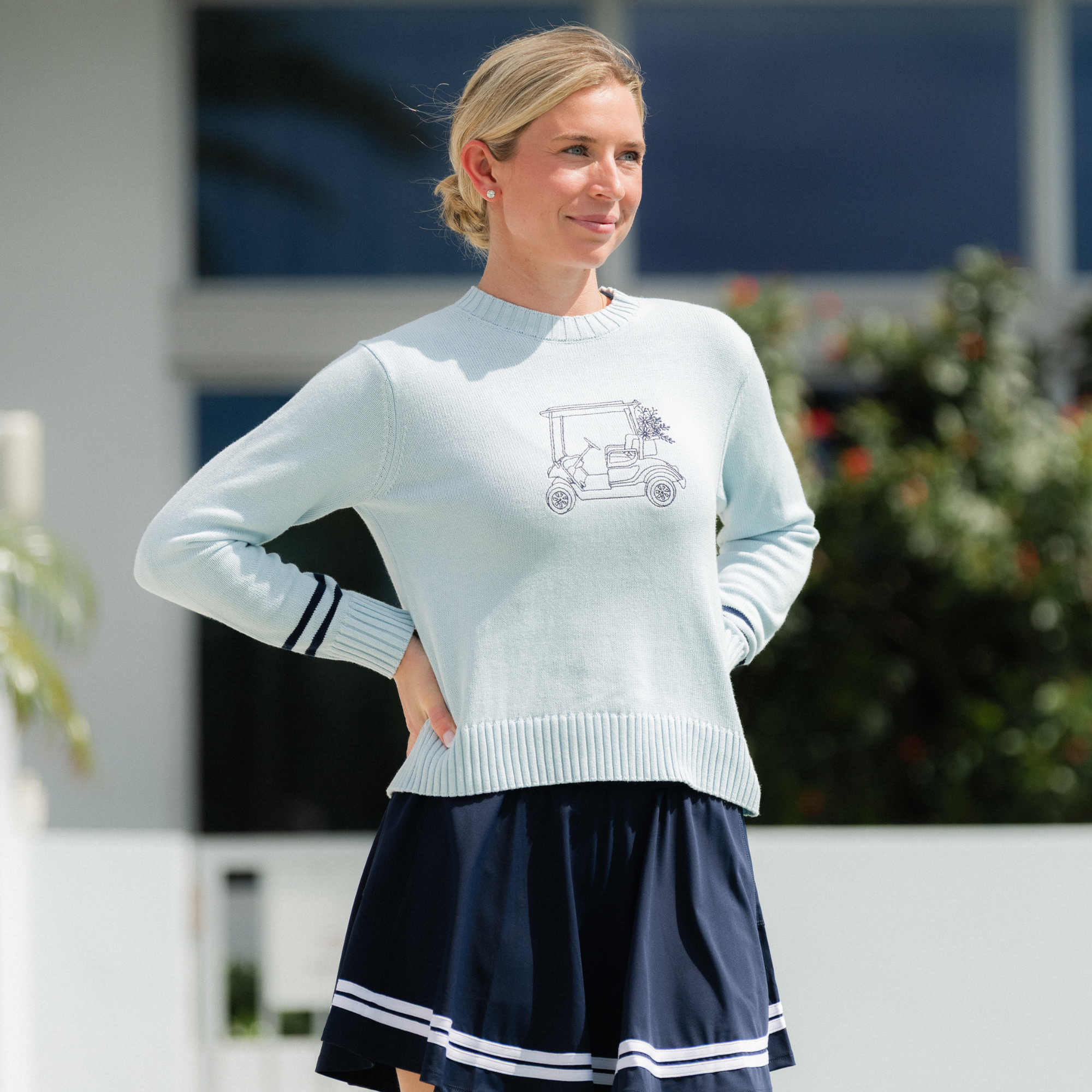 Woman wearing a light blue sweater with a graphic design and a navy skirt outdoors.