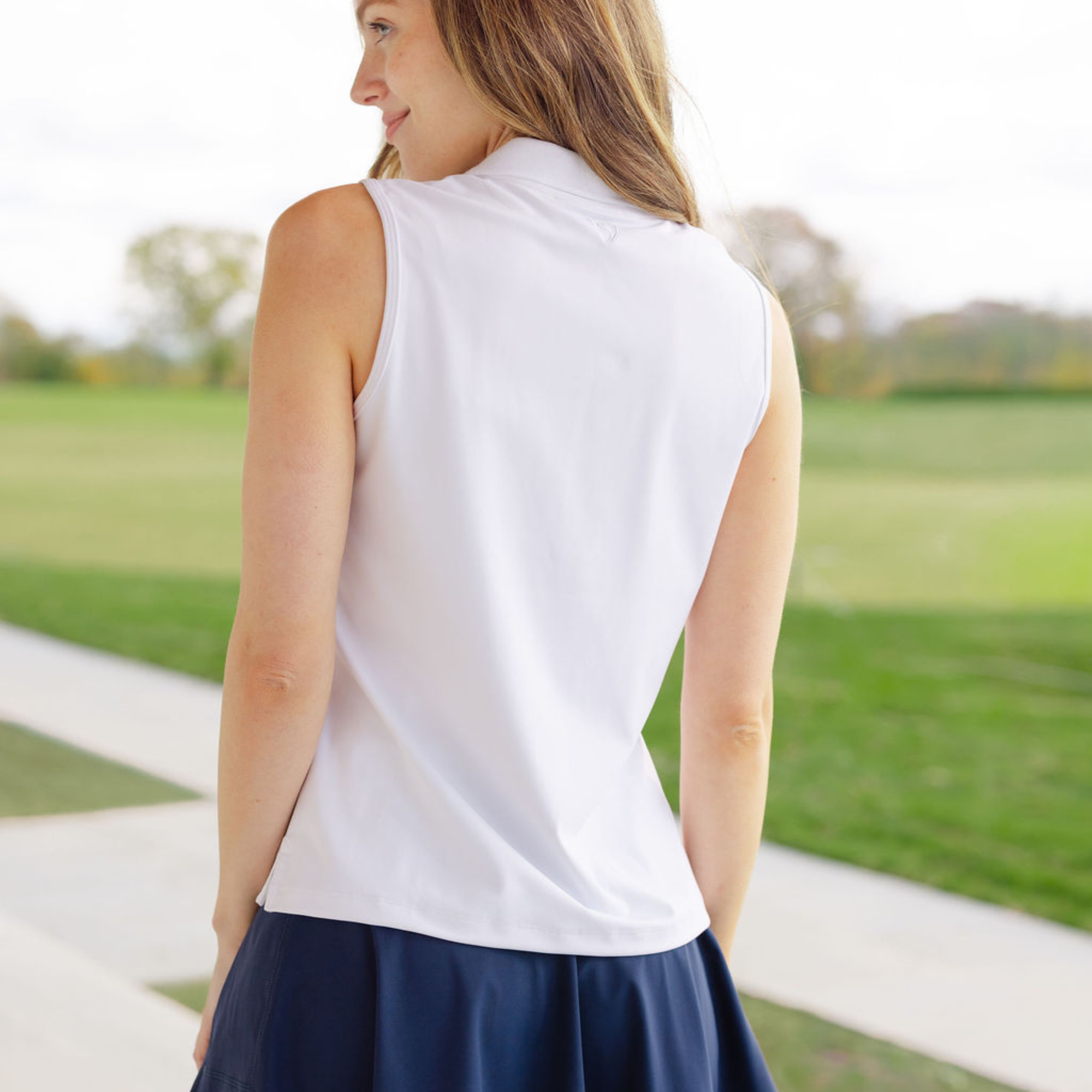 Woman wearing a white sleeveless top and navy skirt standing on a path with grass and trees in the background.