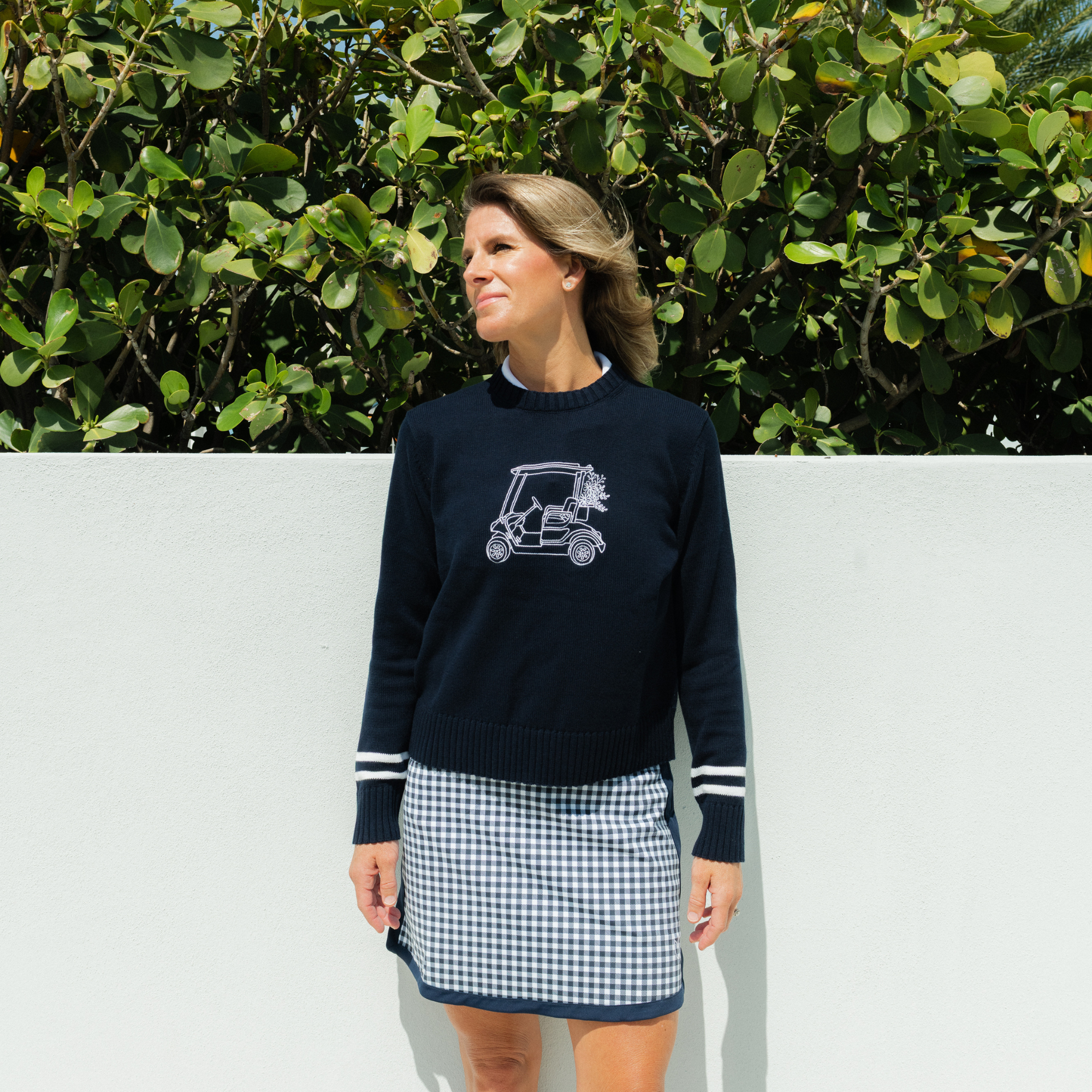 Woman wearing a navy sweater with a graphic design and a checkered skirt against a white wall and green foliage background.