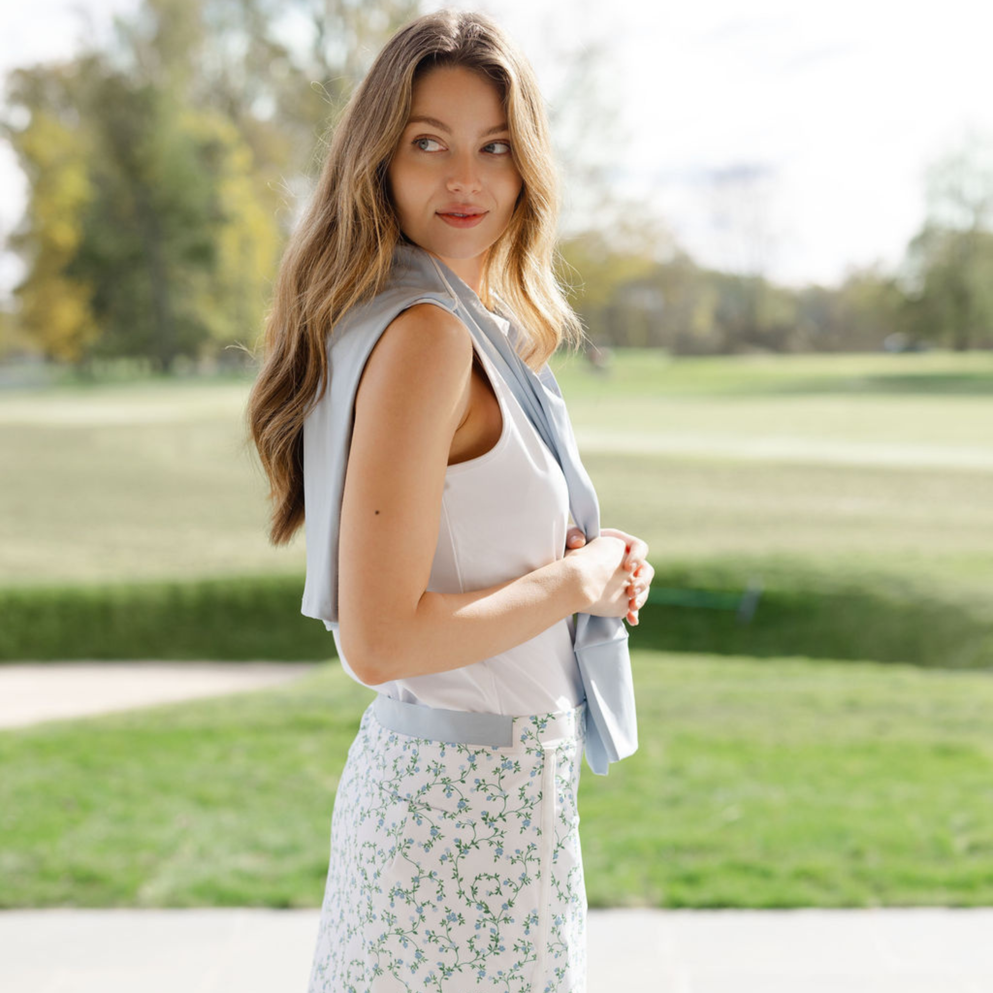 Woman standing outdoors in a park wearing a light blue sleeveless jacket over a white top and floral skirt.