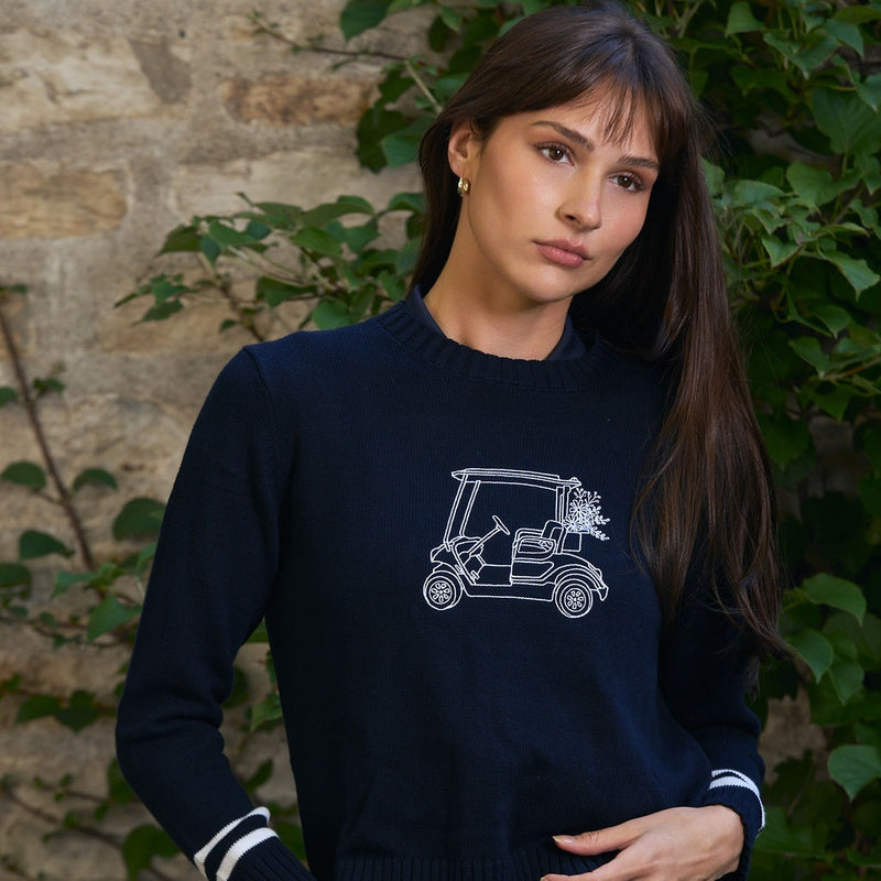 Woman with long dark hair and bangs in a navy crewneck sweater with a white golf cart graphic and striped cuffs, standing in front of a stone wall and green ivy