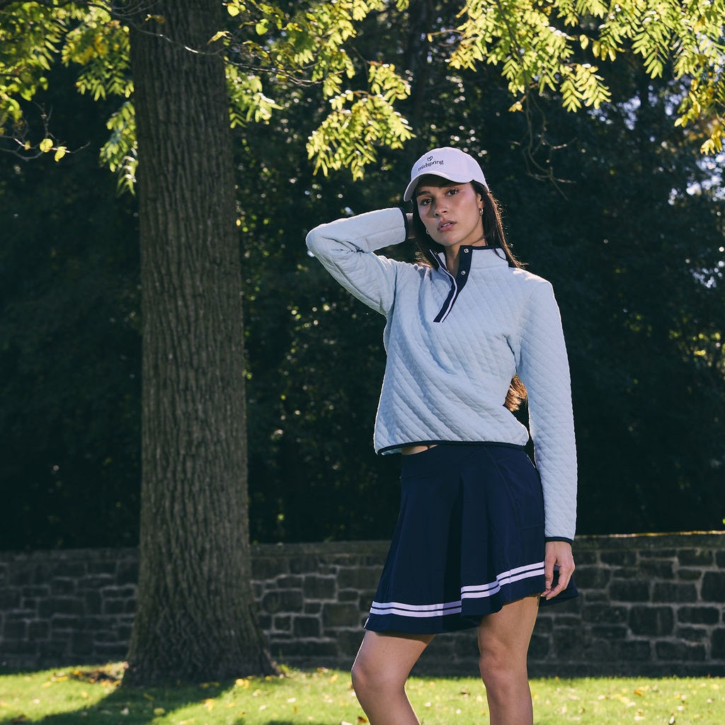 Woman in a white cap with one hand behind her head, wearing a light blue quilted pullover and a navy pleated skirt with white stripes, standing on grass in front of a large tree and a low stone wall