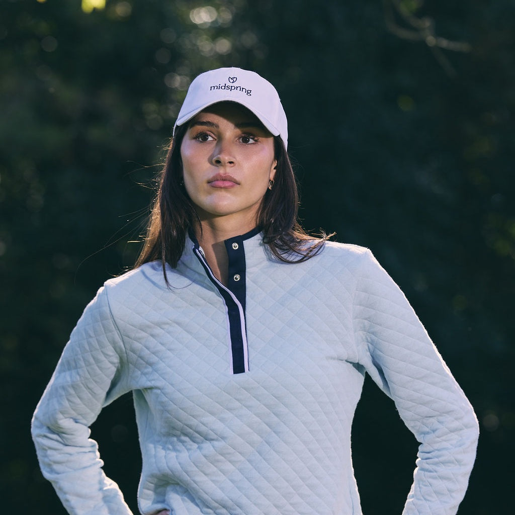 Close-up of a woman in a white cap and a light blue quilted pullover with a navy snap placket, hands on her hips, against a dark blurred green background