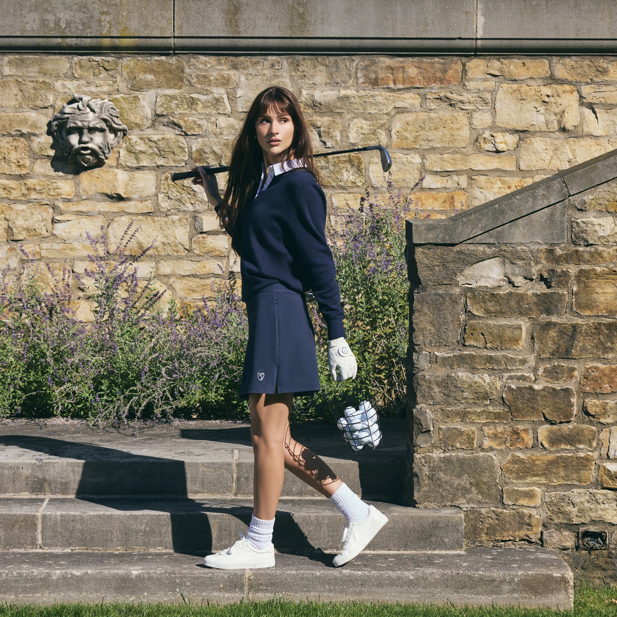 Woman in a navy half-zip pullover and navy pleated golf skirt carrying a golf club over her shoulder as she walks down garden stone steps past a stone wall with a carved face.