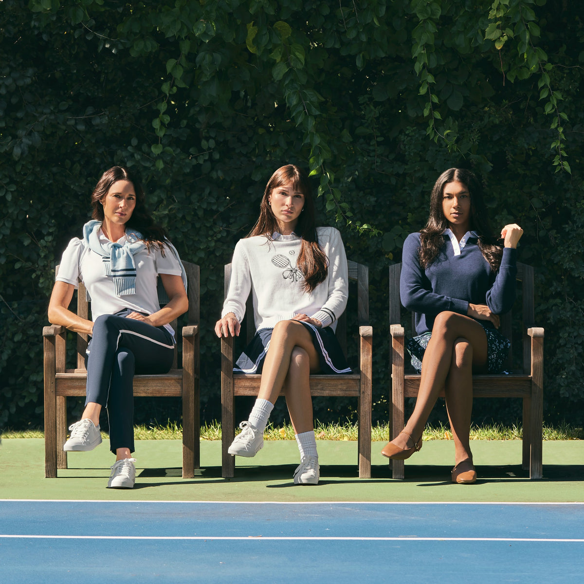 Three women seated side by side in wooden chairs on a blue tennis court in front of a leafy hedge, wearing white, cream and navy tops with navy pants and skirts.